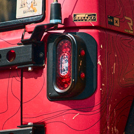 Close-up view of a Tail Gunner Tail Light installed on a red Jeep JK. The tail light features a sleek, oval design with a black aluminum housing and bright red LED lights, providing enhanced visibility. The light assembly includes two smaller red side markers integrated into the design. The durable aluminum construction offers resistance to off-road wear and tear while complementing the rugged appearance of the Jeep.