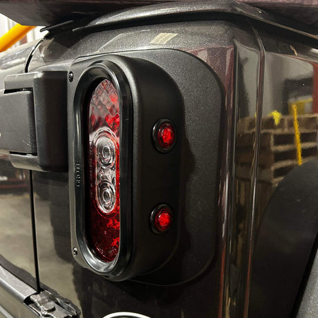 Close-up of a Tail Gunner Tail Light installed on a black Jeep JK. The oval-shaped tail light is housed in a sturdy black aluminum casing, featuring bright red LEDs for the main light and two additional red side markers. The durable and stylish design enhances both visibility and the vehicle's overall aesthetic, suitable for off-road conditions.
