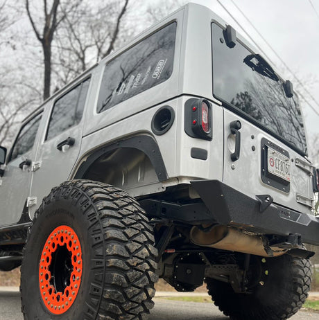 A silver Jeep JK shown from the rear angle, equipped with Tail Gunner Tail Lights featuring red lenses and integrated side marker lights. The vehicle also displays aggressive off-road tires with orange beadlocks, an aftermarket bumper, and raw aluminum fender flares, highlighting its rugged customization. The background includes trees and a cloudy sky, emphasizing the Jeep's outdoor-ready design.