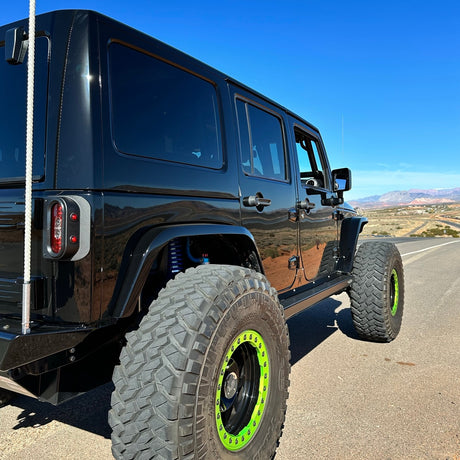 A black Jeep JK photographed from the rear-side angle, featuring Tail Gunner Tail Lights with vibrant red lenses and built-in side markers. The Jeep is outfitted with oversized off-road tires, black wheels, and bright green beadlocks. Custom fender flares and a rear bumper complete the rugged look. The background displays an open road and scenic desert mountains under a clear blue sky.