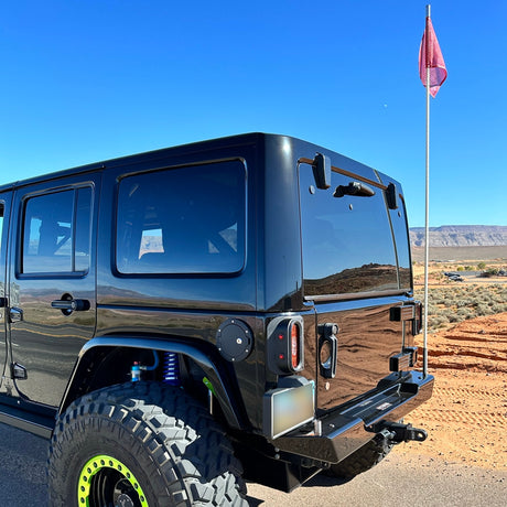 Rear-side view of a black Jeep JK equipped with Tail Gunner Tail Lights featuring red lenses and integrated side markers. The vehicle has oversized off-road tires, black wheels with bright green beadlocks, and a custom rear bumper. A safety whip with a red flag is mounted on the rear, while the desert landscape and clear blue sky provide a scenic backdrop.