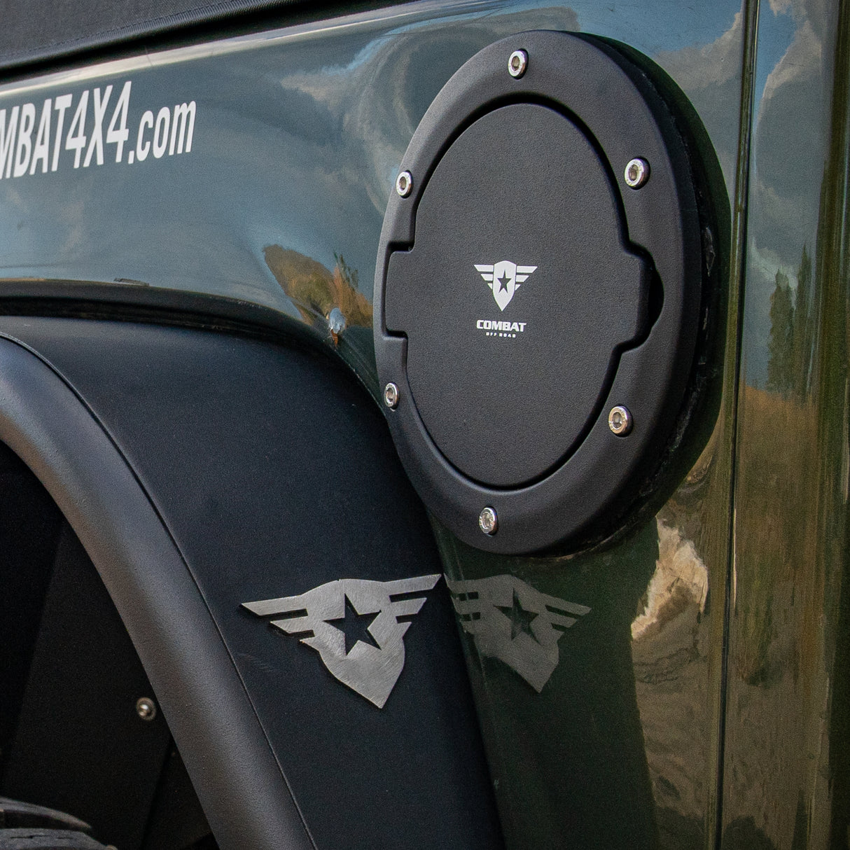 A black Combat Off Road fuel door cover installed on a Jeep JK, featuring the Combat Off Road logo in white at the center. The cover is secured with visible stainless steel bolts for a rugged appearance and provides a sleek, functional upgrade to the fuel compartment. The Jeep's green exterior and fender are also visible in the background.