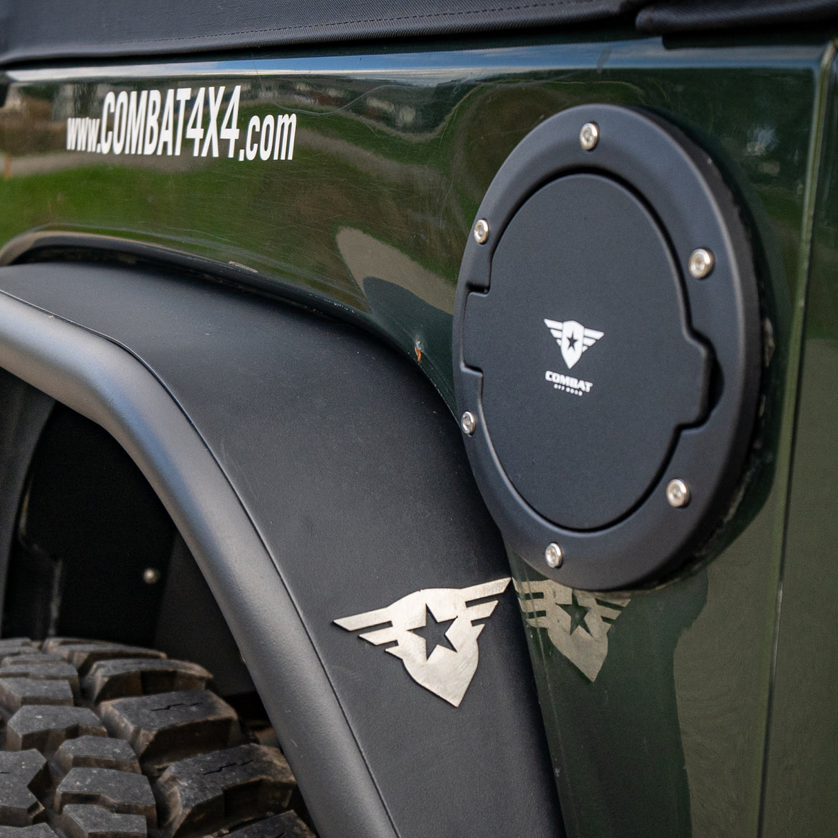 A black Combat Off Road fuel door cover installed on a Jeep JK, featuring a textured finish, stainless steel bolt accents, and the Combat Off Road logo in white at the center. The Jeep’s green exterior and fender are visible, along with a polished Combat Off Road emblem below the fuel door and the website URL "www.COMBAT4X4.com" on the upper body panel. Designed to enhance both function and style for off-road enthusiasts.